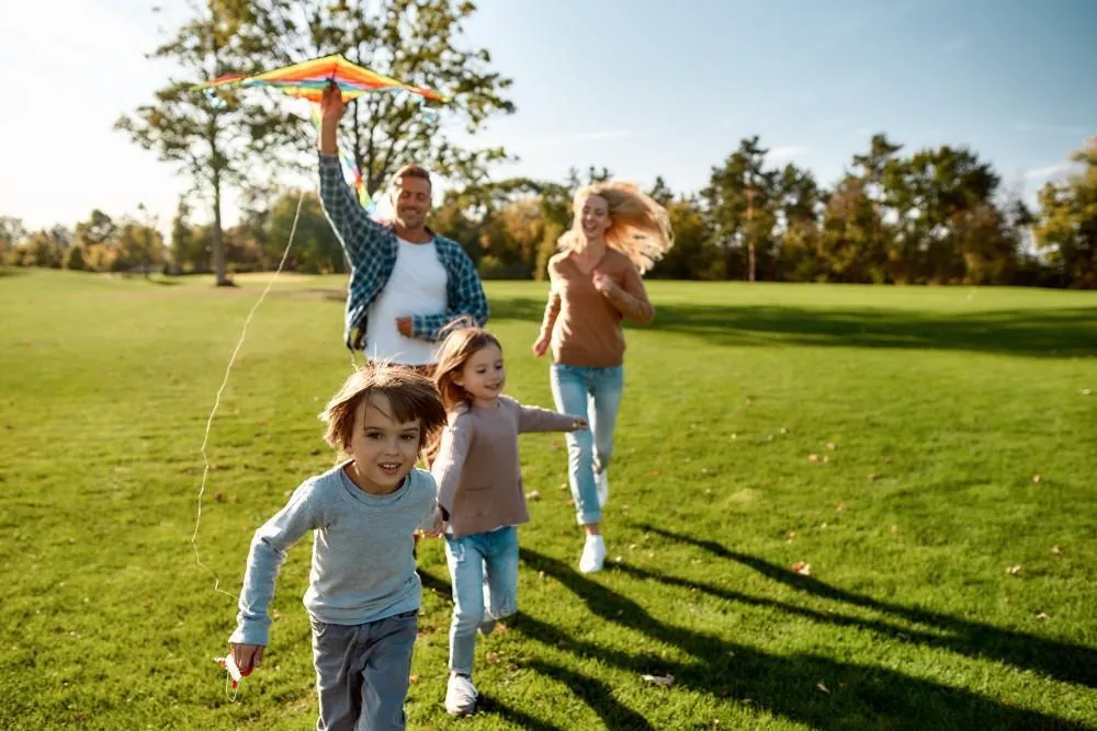 A family with two young children runs across a sunny park lawn, flying a rainbow kite.