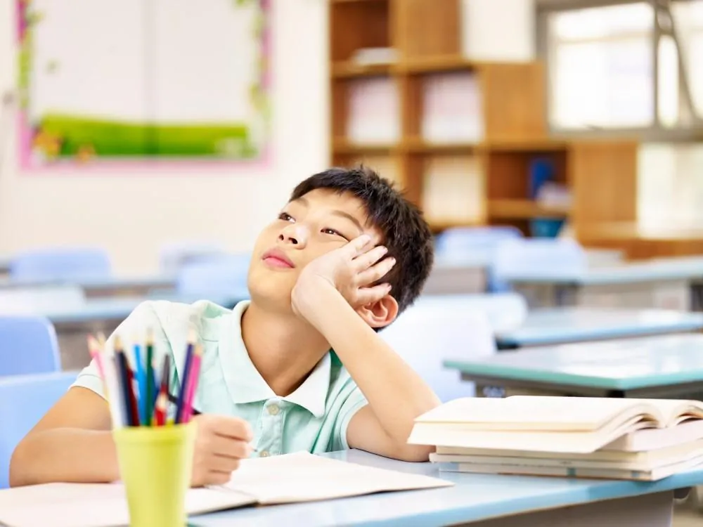 A school-age boy rests his head on his hand and gazes upward, distracted from the open books on his classroom desk.