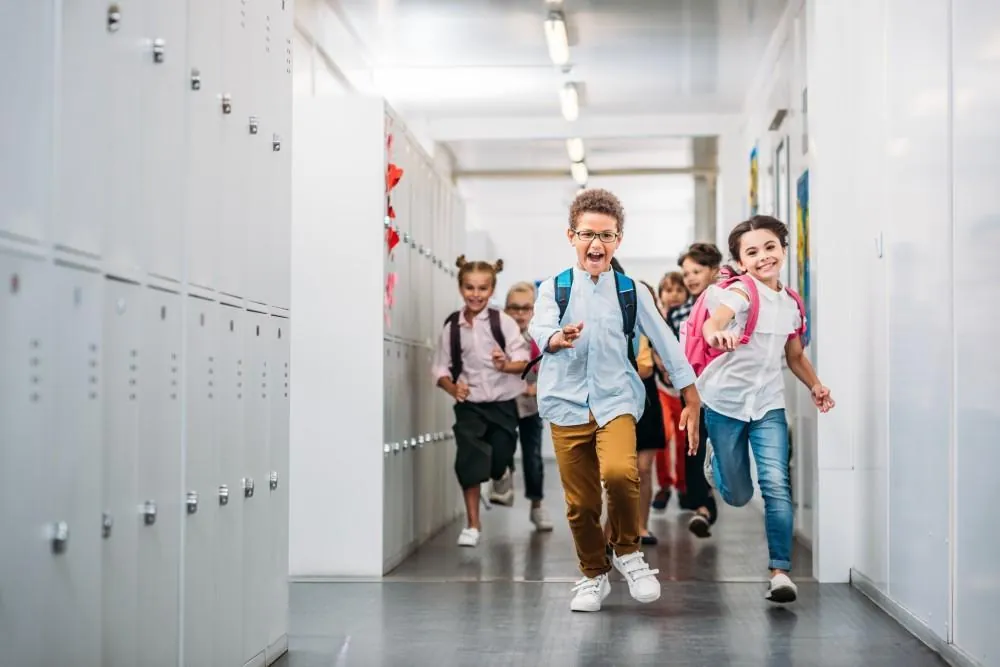 Children wearing backpacks run down a school hallway lined with lockers.