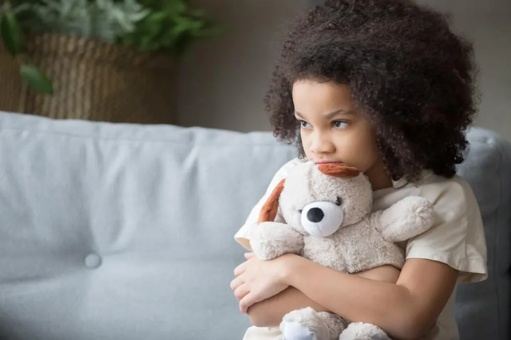 A young girl sits on a sofa hugging a stuffed toy, looking away with a downcast expression.