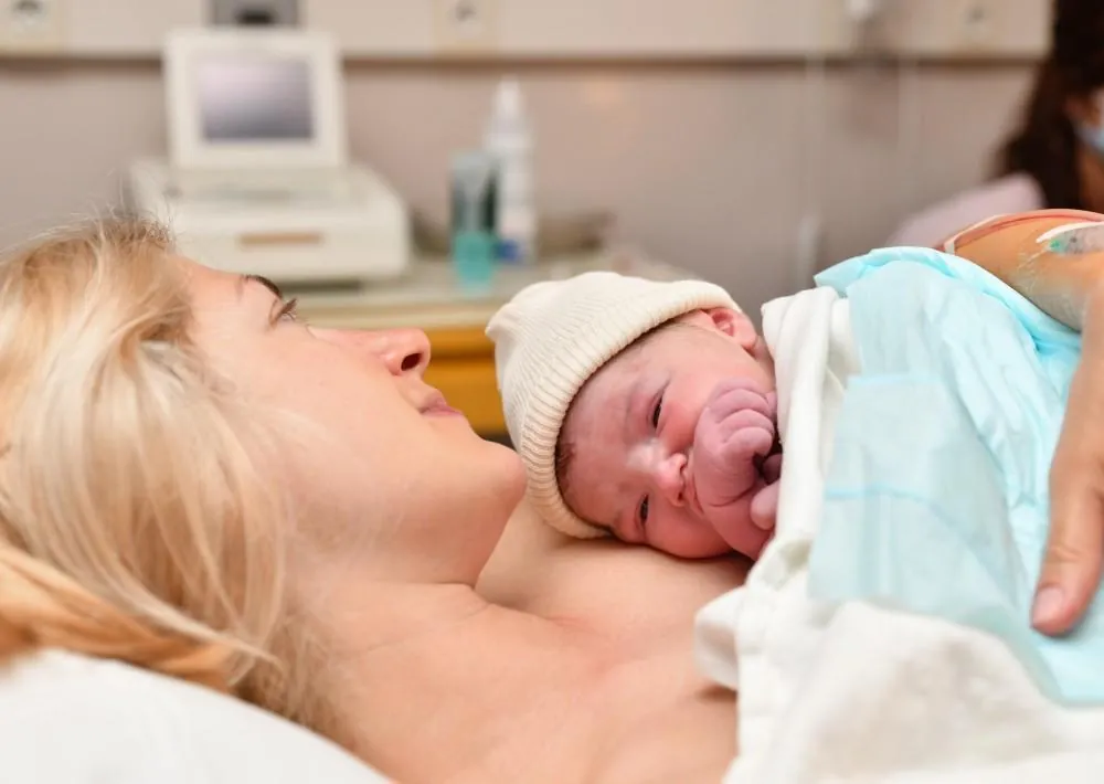 A mother rests in a hospital bed holding her newborn skin to skin, the baby wearing a knitted cap.