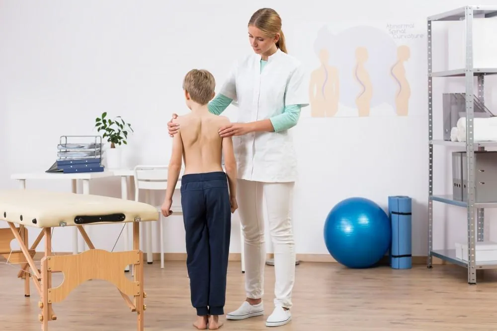 A clinician examines a boy's back during a physical exam, beside a wall chart showing abnormal spinal curvature.