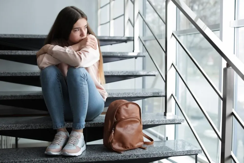 A teenage girl sits alone on a stairwell with her arms wrapped around her knees, her backpack on the step beside her.