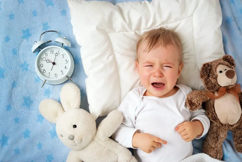 A baby lies crying on a blue blanket between two stuffed animals and a small alarm clock.