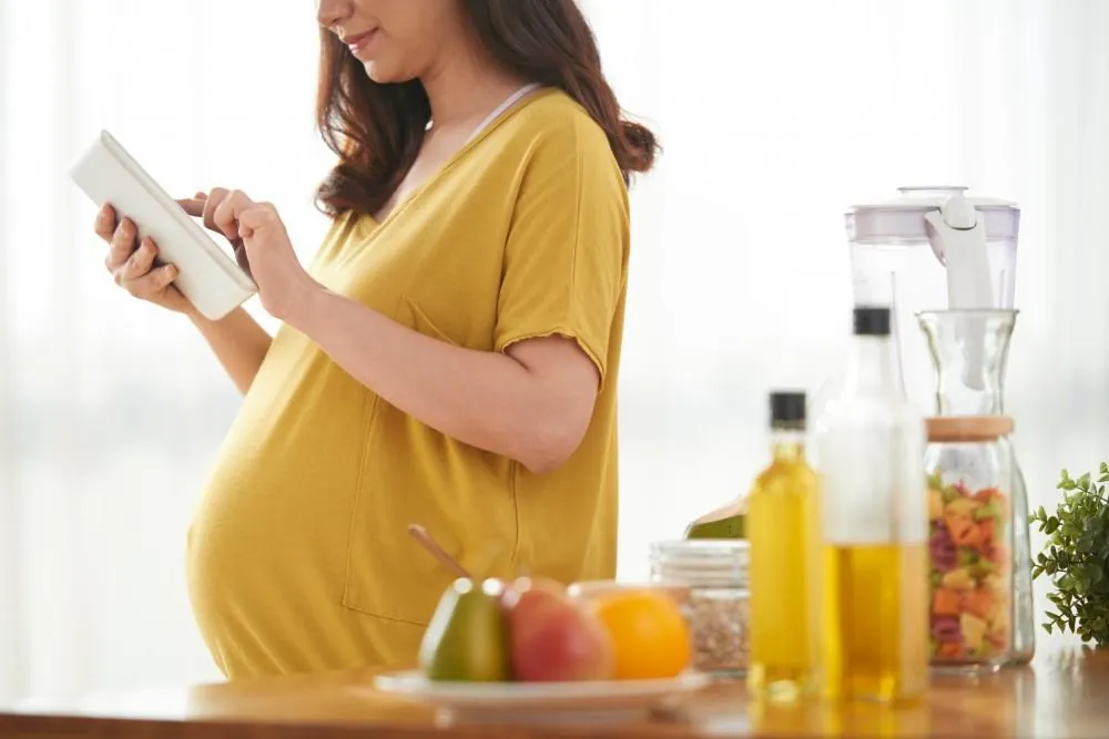 A pregnant woman reads a tablet in a kitchen, with fresh fruit, cooking oils, and a jar of pasta on the counter.