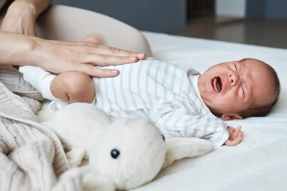 A parent rests a hand on a crying newborn's stomach as the baby lies on a bed beside a soft toy.
