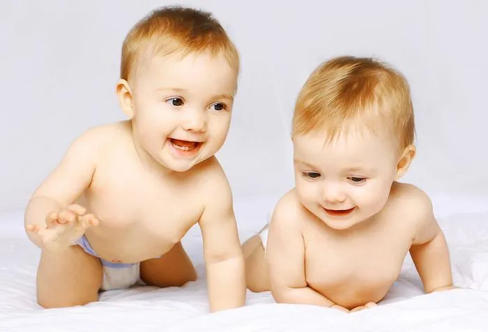 Two smiling babies in diapers lie side by side on a white blanket.