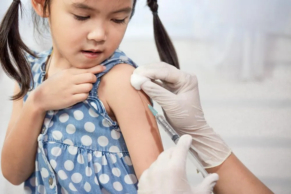 A young girl in a polka-dot dress watches as a gloved clinician gives her a vaccination in the arm.