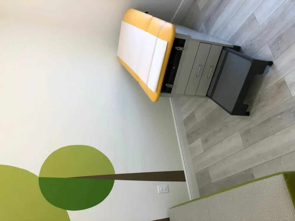 Exam room with a yellow examination table, step stool, and a painted tree mural on the wall.