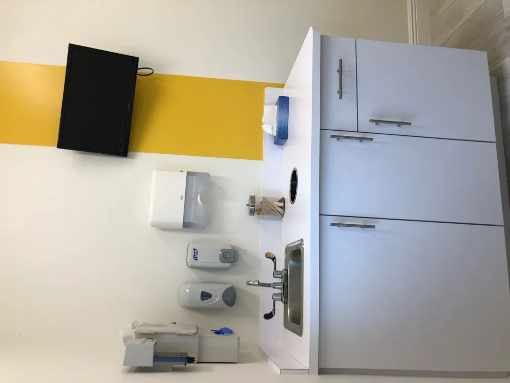 Exam room counter with a sink, soap and sanitiser dispensers, and a wall-mounted television.