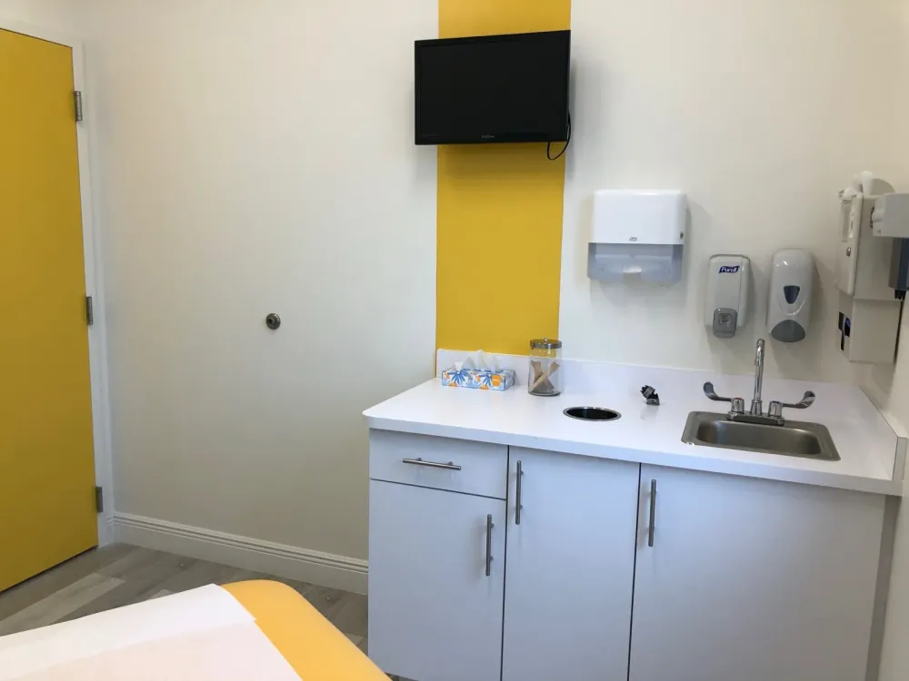 Exam room with a sink counter, wall-mounted television, and a yellow door.