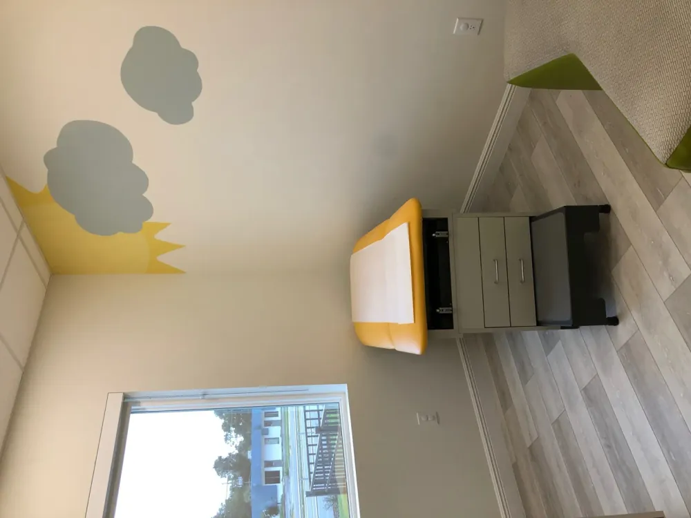 Exam room with a large window, yellow exam table, and a painted sun and clouds on the wall.