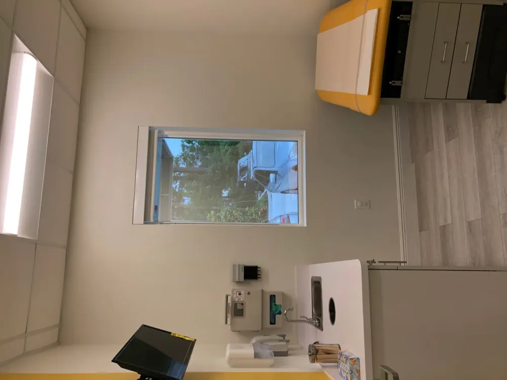Exam room with a window, sink counter, wall-mounted television, and a yellow exam table.
