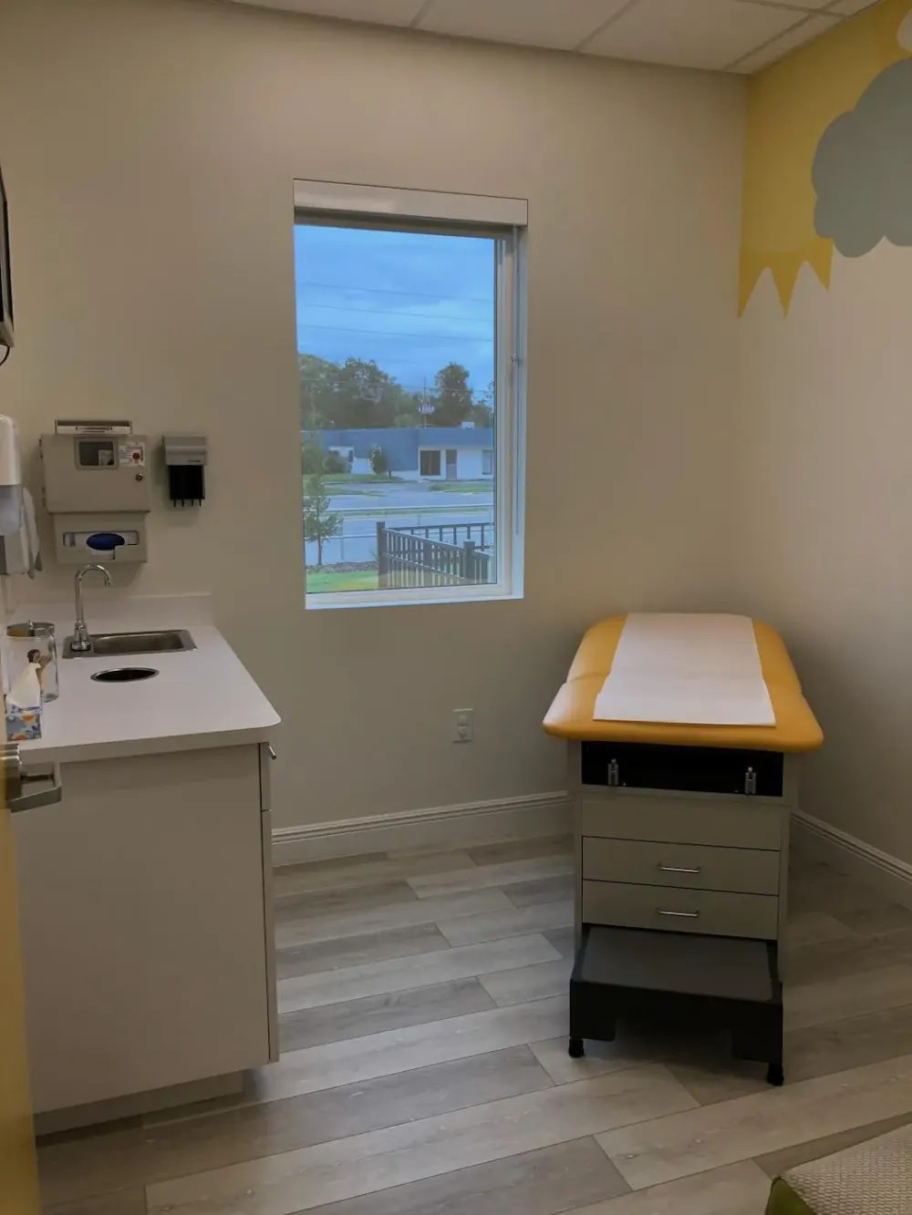 Exam room with a sink counter, window, and a yellow exam table beneath a cloud mural.
