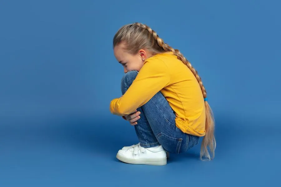 A teenager sitting alone on a sofa, looking down, with their arms folded.